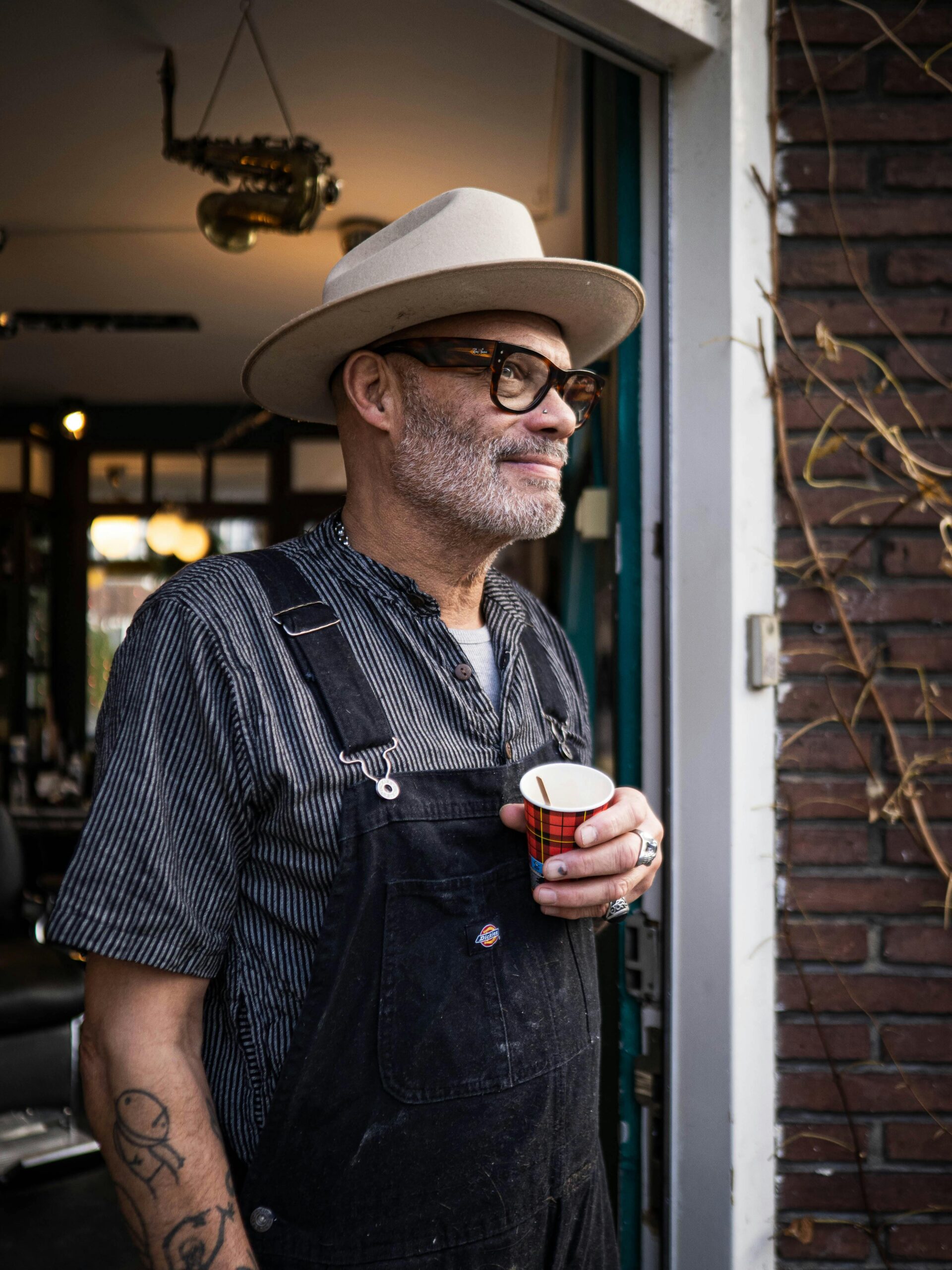 Man standing in doorway with coffee cup and wearing hat and overalls in urban setting.
